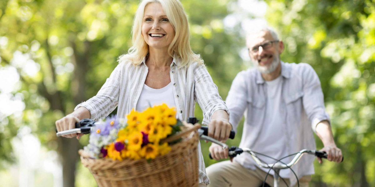 Active Retirement. Beautiful Senior Woman Riding Bycicles With Her Husband In Park, Happy Mature Man And Woman Enjoying Outdoor Leisure, Making Bike Ride And Having Fun Together Outside, Free Space