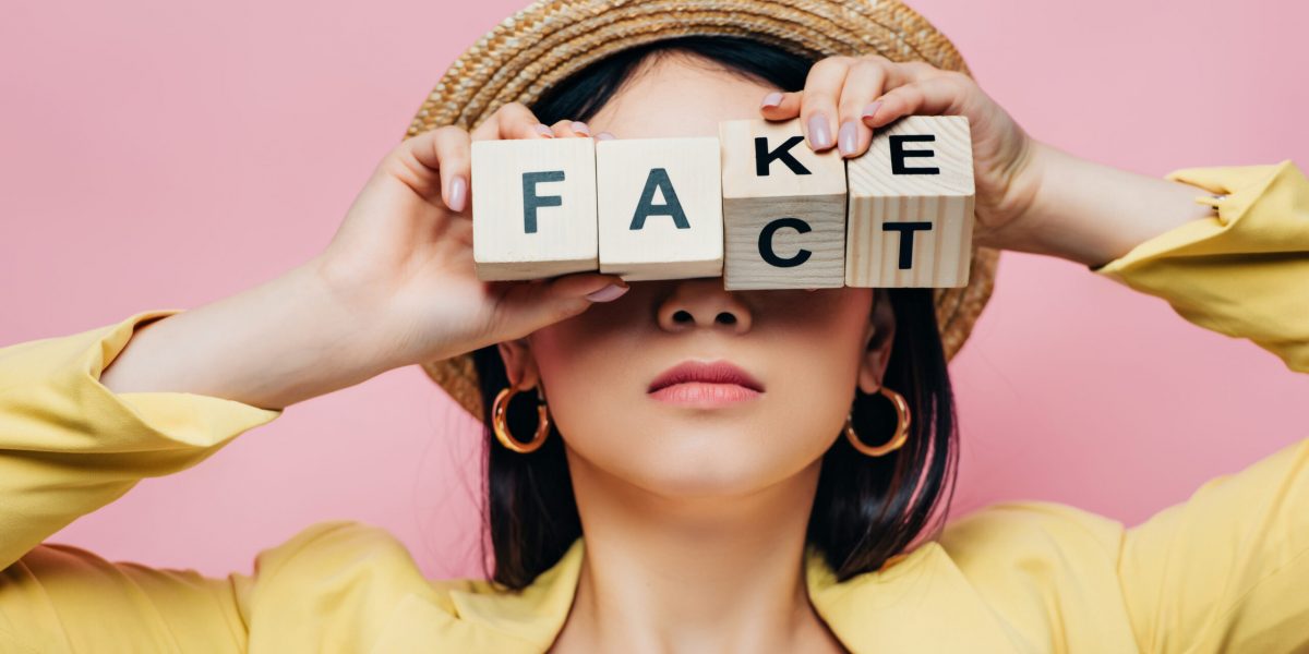 asian woman holding wooden cubes in front of face with fake and fact lettering isolated on pink
