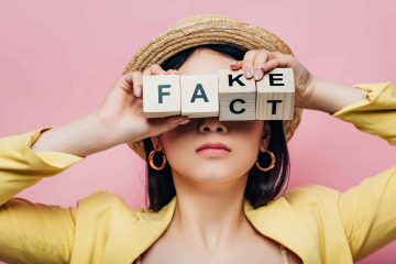 asian woman holding wooden cubes in front of face with fake and fact lettering isolated on pink