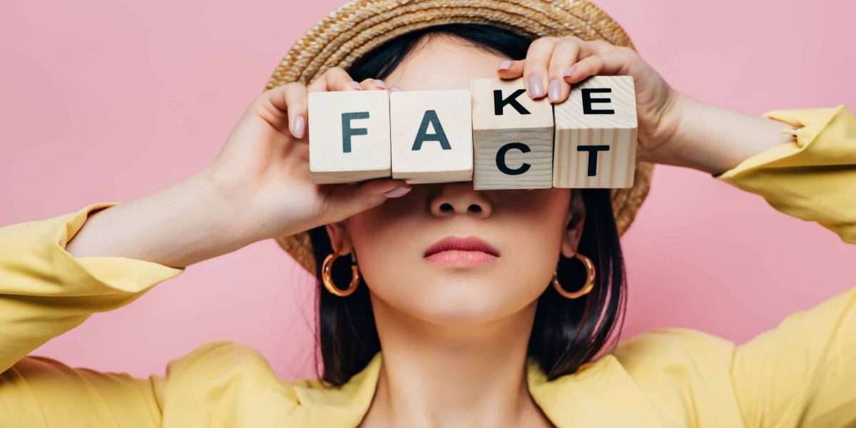 asian woman holding wooden cubes in front of face with fake and fact lettering isolated on pink