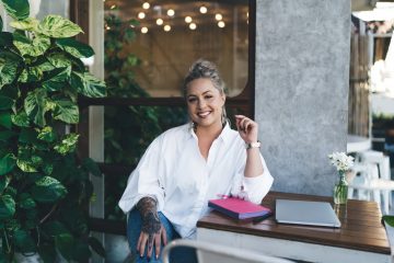 Portrait of happy female writer dressed in casual white shirt smiling at camera during daytime weekend in cafe, cheerful Caucasian student with education textbook for planning posing at table