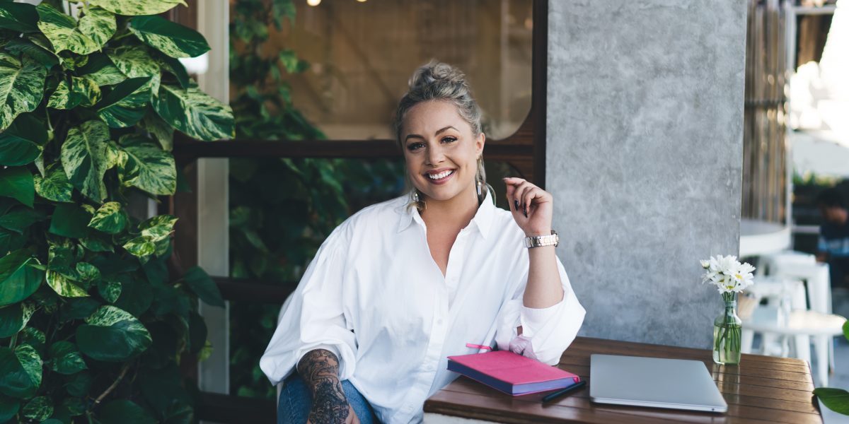 Portrait of happy female writer dressed in casual white shirt smiling at camera during daytime weekend in cafe, cheerful Caucasian student with education textbook for planning posing at table