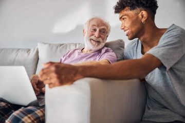 Merry senior Caucasian man with a laptop looking at a young curly-haired African American caregiver