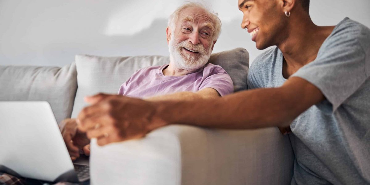 Merry senior Caucasian man with a laptop looking at a young curly-haired African American caregiver