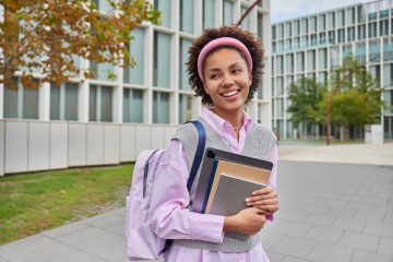 Cheerful schoolgirl carries rucksack notepads and digital tablet looks into distance with happy expression walks in campus during daytime. Female student returns from university. Education concept