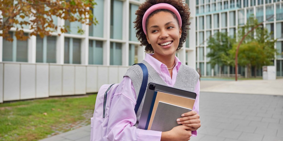 Cheerful schoolgirl carries rucksack notepads and digital tablet looks into distance with happy expression walks in campus during daytime. Female student returns from university. Education concept