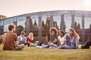 Group Of University Or College Students Sit On Grass Outdoors On Campus Talking And Working