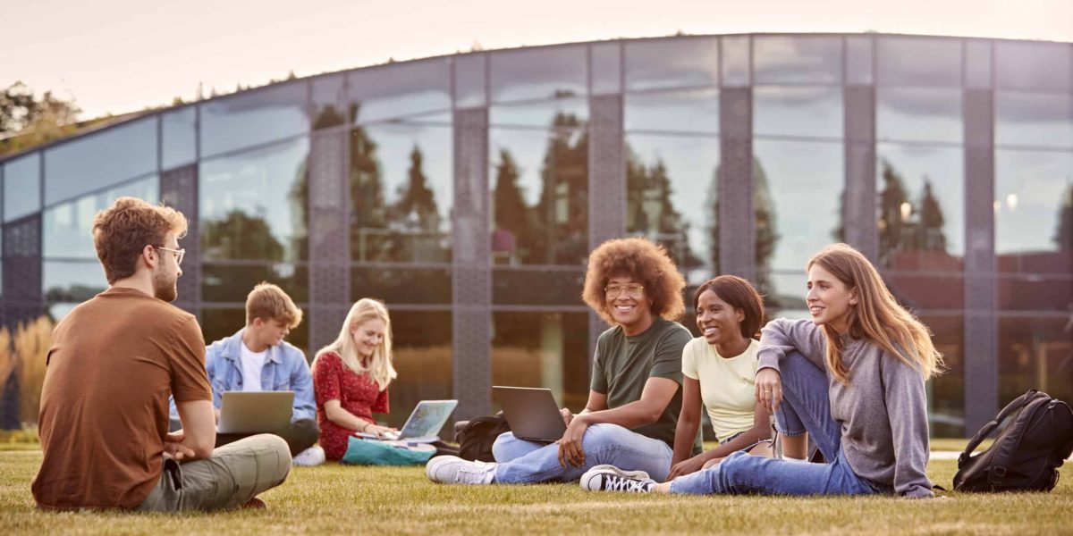 Group Of University Or College Students Sit On Grass Outdoors On Campus Talking And Working