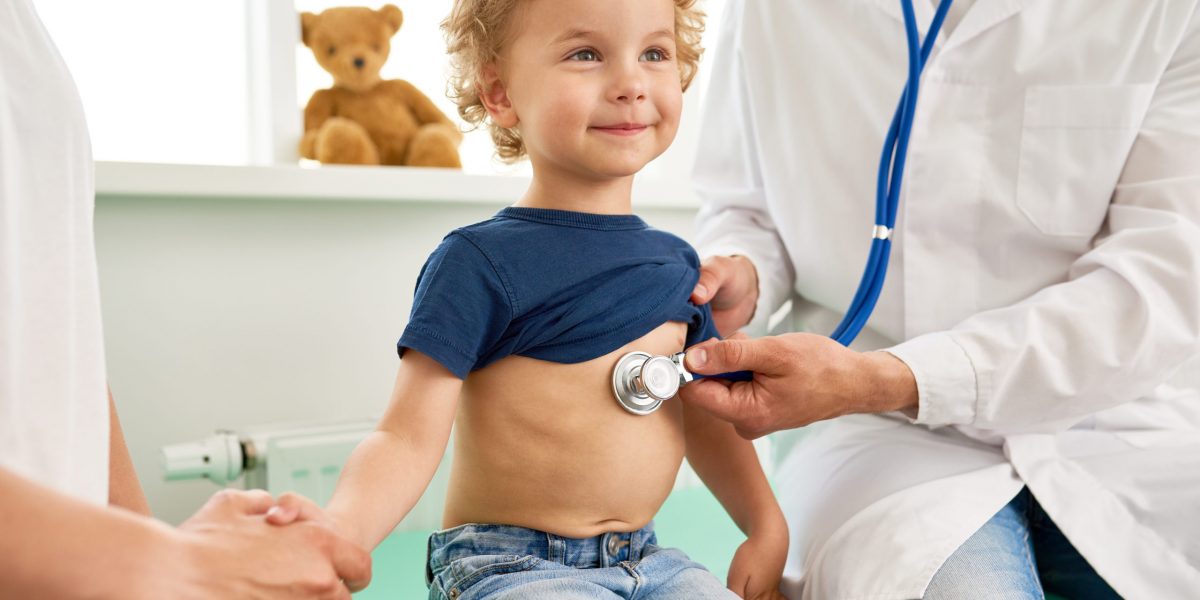 Portrait of adorable little boy visiting doctor, looking brave and smiling, holding mothers hand while pediatrician listening to heartbeat with stethoscope