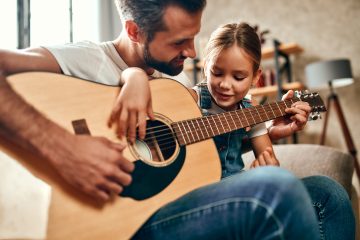 Happy dad teaches his cute daughter to play the guitar while sitting on the sofa in the living room at home. Happy Father's Day.
