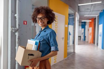 Happy Afro American lady carrying a box of documents down the hallway in the office