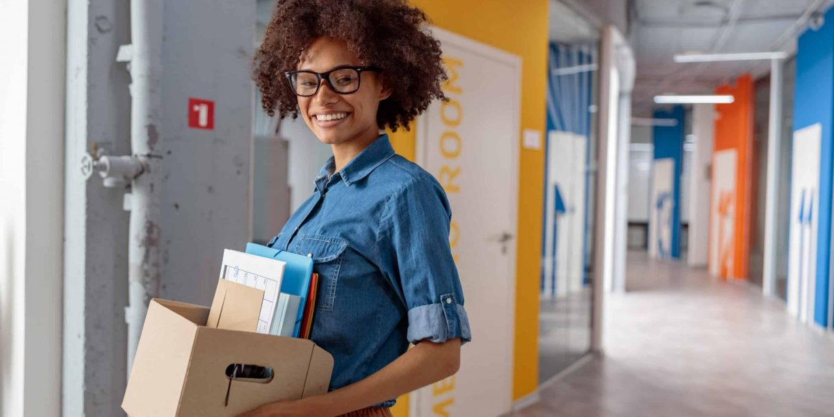 Happy Afro American lady carrying a box of documents down the hallway in the office