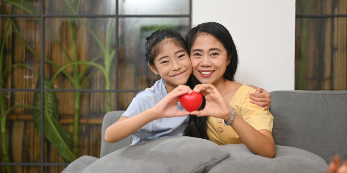 Little girl and mother holding heart shape and smiling at camera. Life insurance, love and support in family relationships concept.