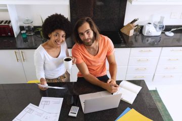 Portrait of happy diverse couple sitting in kitchen using laptop and paying bills. staying at home in isolation during quarantine lockdown.