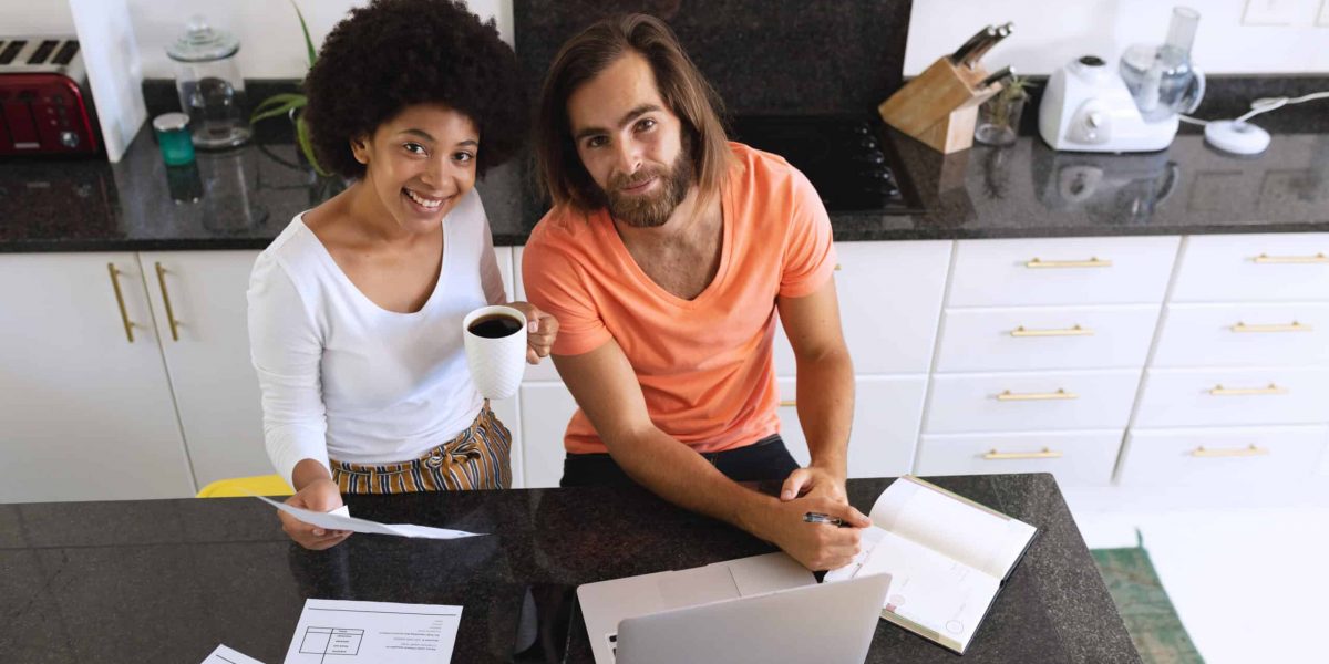 Portrait of happy diverse couple sitting in kitchen using laptop and paying bills. staying at home in isolation during quarantine lockdown.