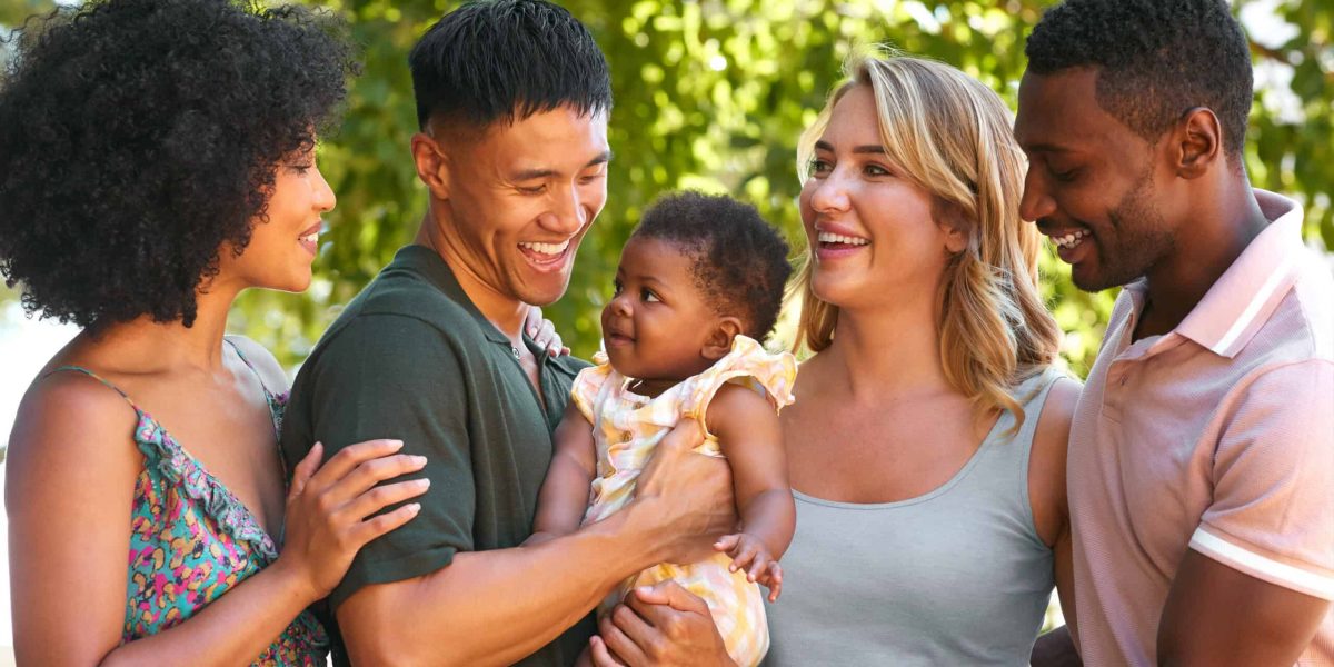 Portrait Of Multi-Racial Family And Friends Hugging Baby Outdoors In Countryside In Group Together