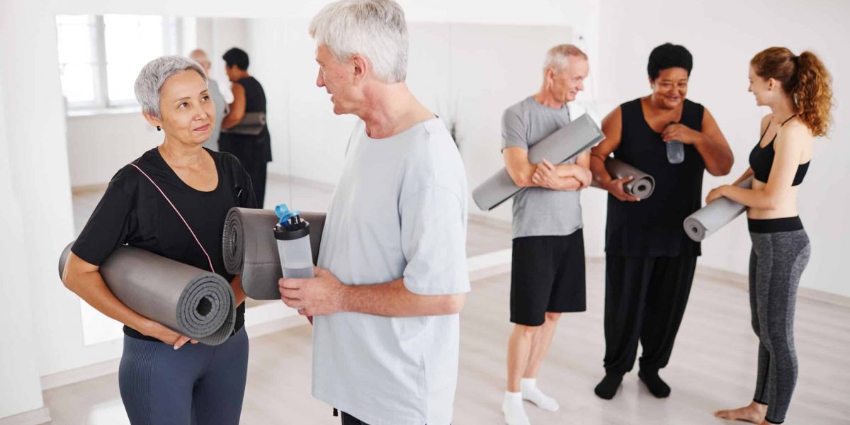 Senior couple with exercise mats talking to each other after yoga class with other people in the background